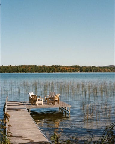 Wooden dock with Adirondack chairs extending into calm lake, forested shoreline and clear sky beyond