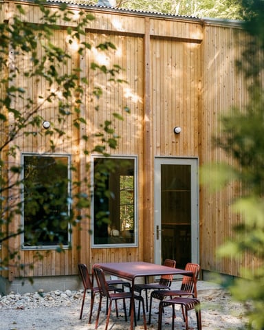 Modern wooden cabin exterior with metal-framed windows and red chairs at a patio table, surrounded by green foliage.