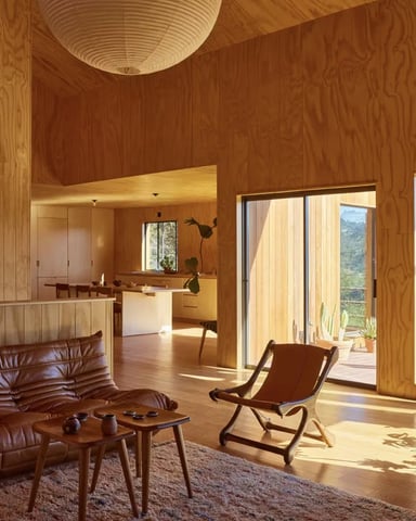Wood-paneled living room with red leather sofa, mid-century chair, and glass doors opening to forested deck with mountain views