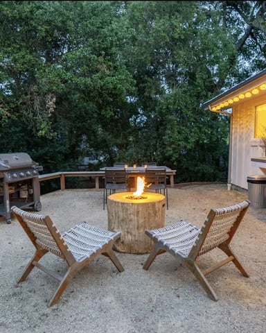 Backyard patio at dusk with fire pit, two lounge chairs, grill, and illuminated modern house surrounded by tall trees