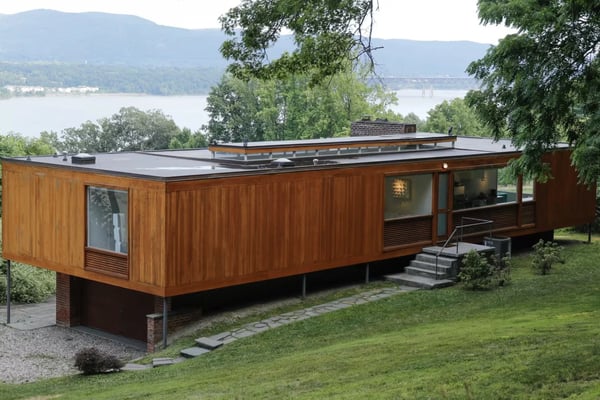 Modern wooden cabin with flat roof on stilts, surrounded by green lawn and trees, overlooking a lake and mountains