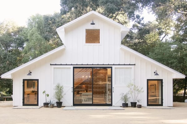 Modern white barn with black-framed glass doors and windows, surrounded by tall trees