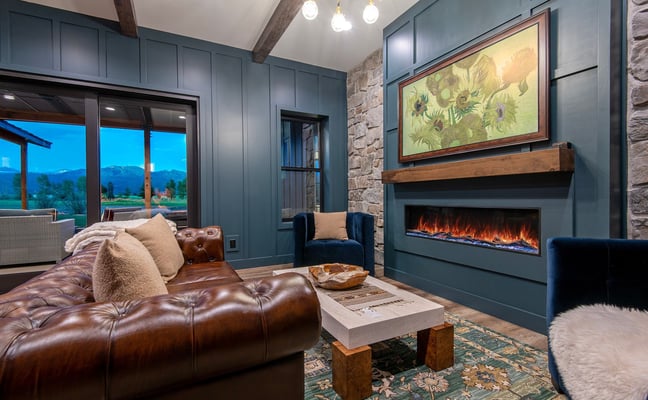 Modern living room with brown leather sofa, electric fireplace, blue-green paneled walls, stone accent wall, and mountain views through glass doors.