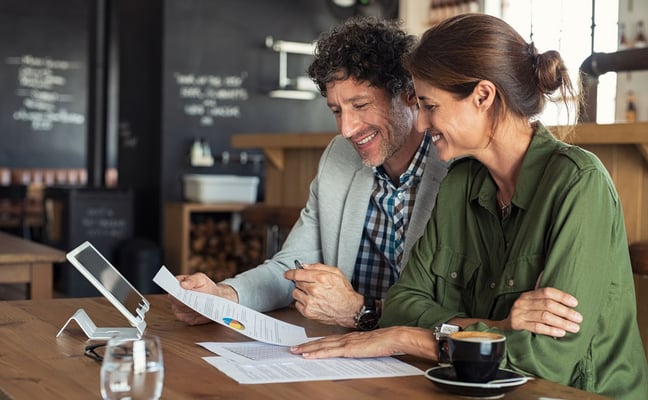 Two people reviewing documents and tablet at wooden table in modern café with dark menu board backdrop