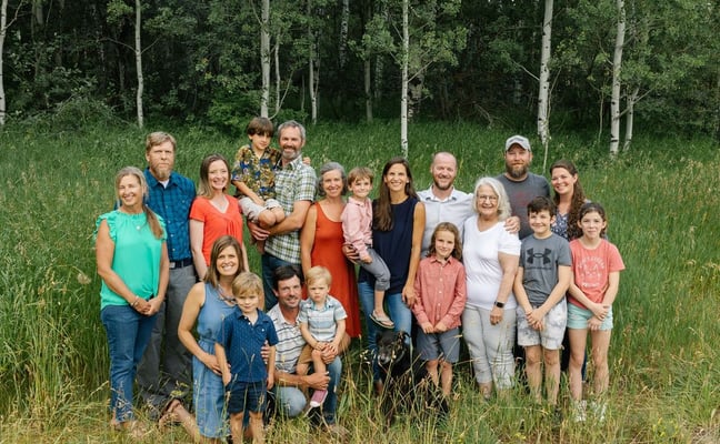 Large family group of adults and children standing together in a grassy field with birch and evergreen trees in the background