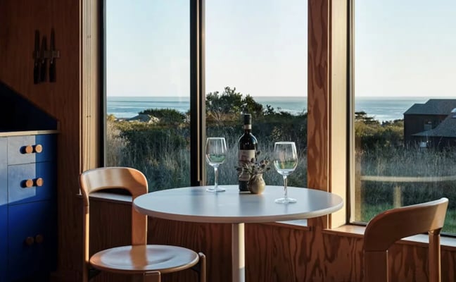 Dining room with round table, wooden chairs, wine bottle and glasses, overlooking coastal landscape through large windows