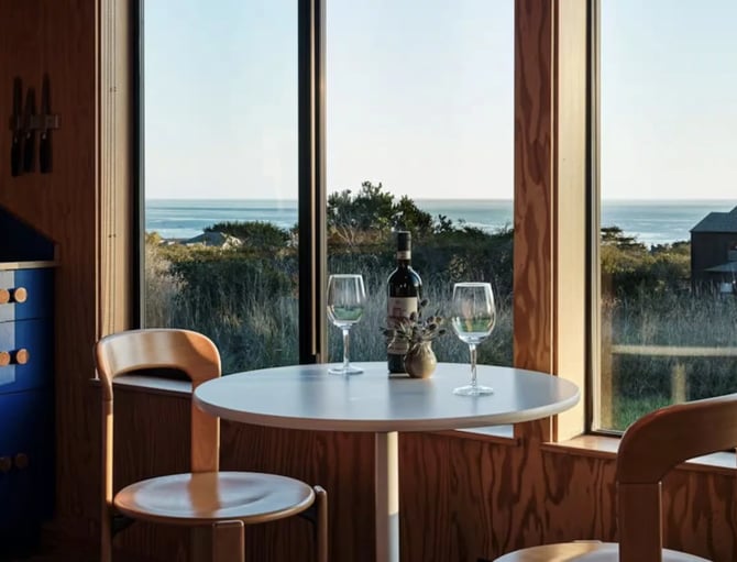 Dining room with round table, wooden chairs, wine bottle and glasses, overlooking coastal landscape and ocean through large windows
